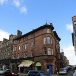 Corner view of a historic red sandstone building on Great Junction Street, Edinburgh, featuring a mix of residential and commercial spaces, with blue sky and clouds in the background.