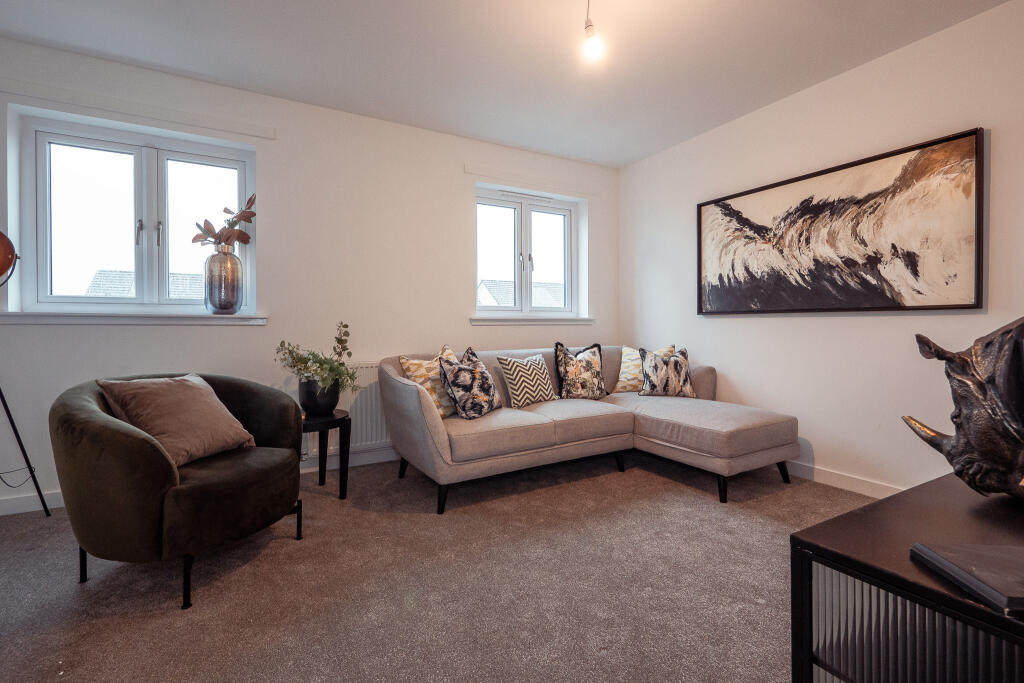 Living room of a 4-bedroom unfurnished terraced house in South Queensferry, featuring a grey sectional sofa with decorative cushions, a green armchair, a modern artwork on the wall, and large windows allowing natural light.