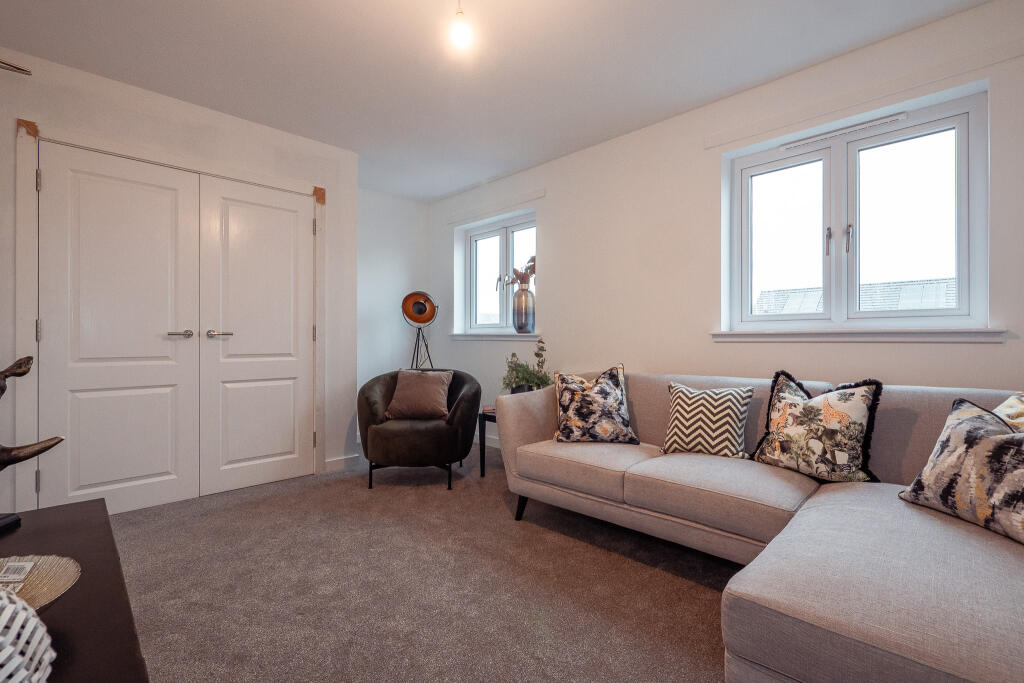 Living room interior of a 4-bedroom unfurnished terraced house in South Queensferry, featuring a grey sectional sofa with decorative cushions, a stylish armchair, and two windows providing natural light, complemented by a modern decorative piece and a carpeted floor.